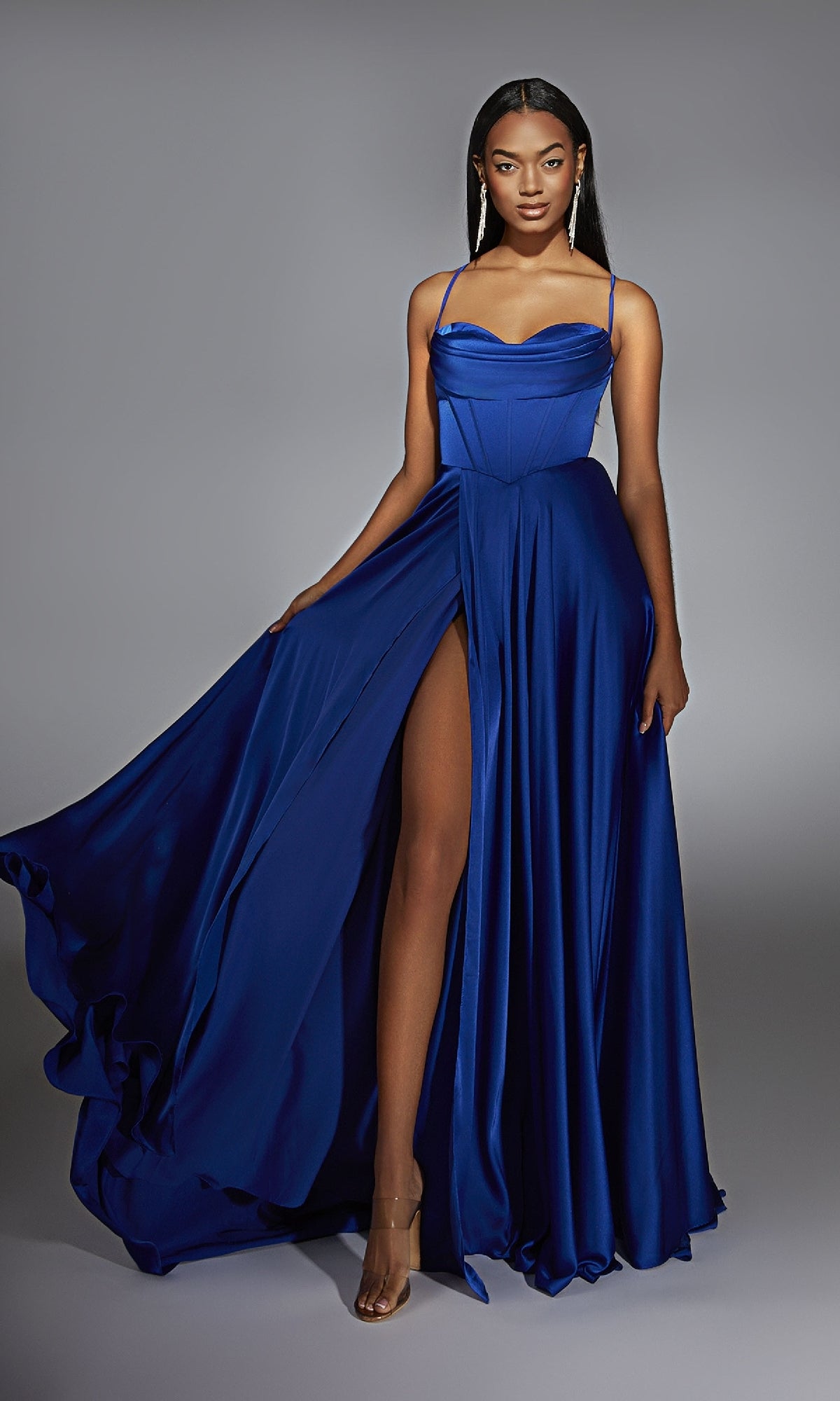 A woman models the Alyce 61883 Long Sweetheart A-Line Prom Dress in royal blue with a floor-length skirt, corset bodice, and high slit, standing against a neutral gray background.